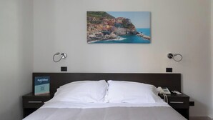 Ein Hotelzimmer mit einem Bett, einem Nachttisch mit Telefon, einem gerahmten Bild einer Küstenstadt und einem Schild, das ein Urlauspaket anzeigt.