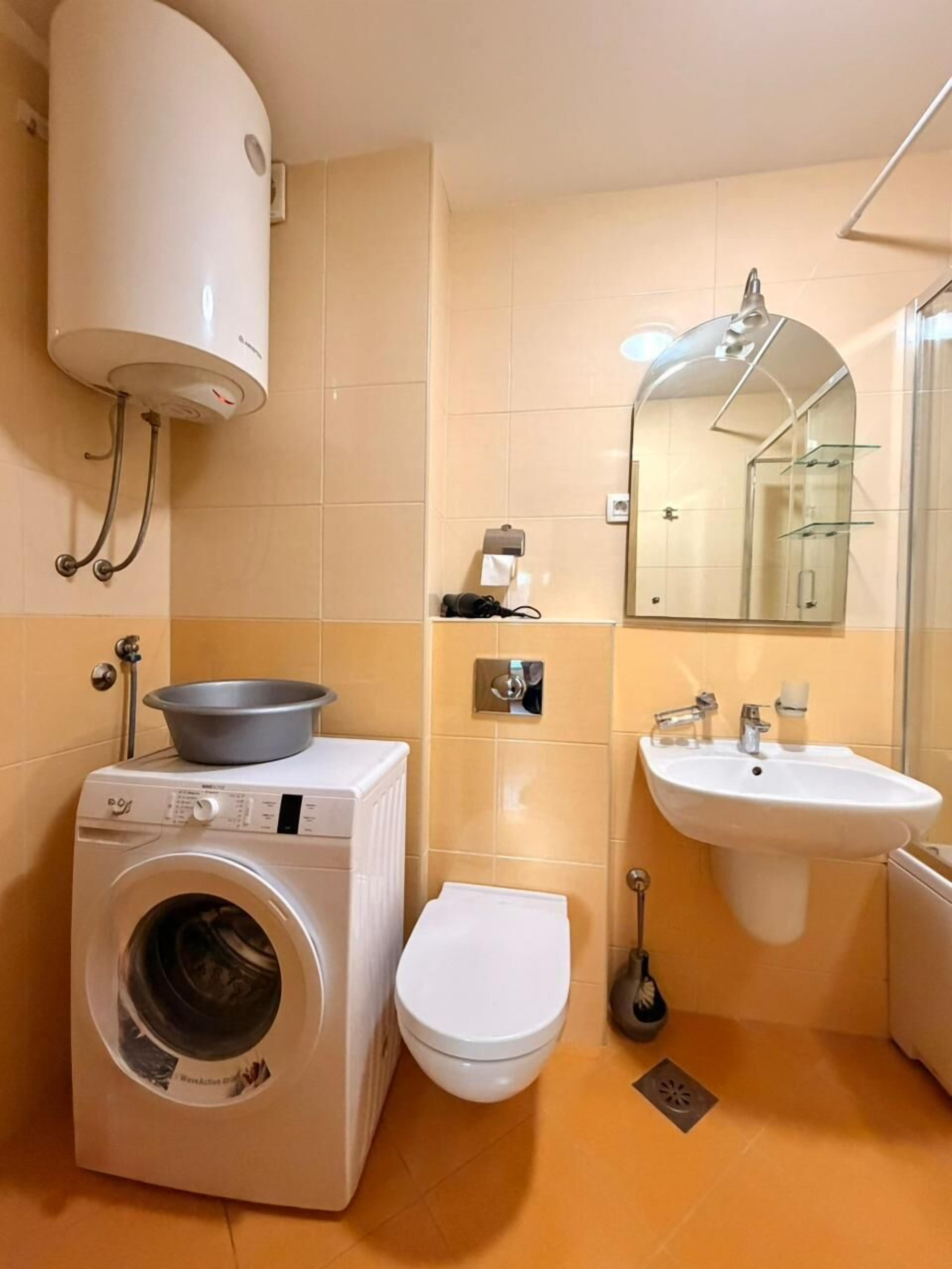 A compact bathroom with a washing machine, toilet, sink, and a mirror with a glass shelf.