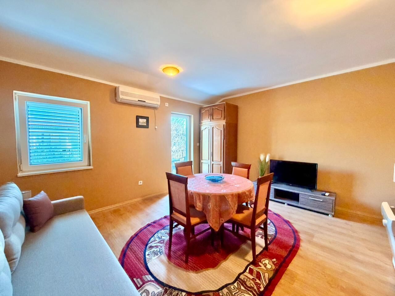 Ein Wohnzimmer mit rundem Tisch, Stühlen, einer Couch, einem Fernseher und einem Fenster mit Jalousien.