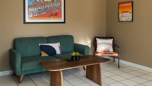 Ein Wohnzimmer mit einem grünen Sofa, einem Holztisch und einem Sessel mit Kissen. An der Wand befinden sich zwei gerahmte Poster.