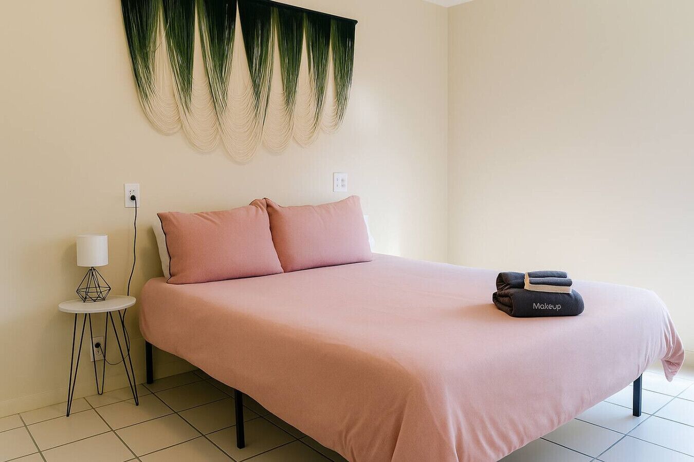 A neatly made bed with pink bedding, a small bedside table with a lamp, and a makeup bag on the bed.