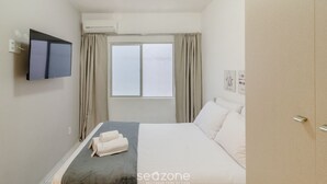 A hotel room with a bed, a flat-screen TV mounted on the wall, an air conditioning unit, a window with curtains, and a wardrobe. - Apt With A/C 350m From the Beach in BC Esf0203 (Balneário Camboriú)