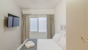 A hotel room with a bed, a flat-screen TV mounted on the wall, an air conditioning unit, a window with curtains, and a wardrobe.