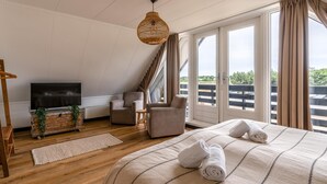 Ein Schlafzimmer mit einem großen Bett, einem Fernseher, zwei Sesseln, einer hölzernen Kommode und einem großen Fenster mit Vorhängen.