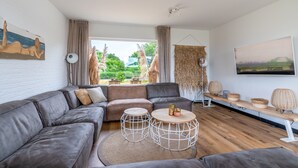 Ein modernes Wohnzimmer mit einem grauen Ecksofa, zwei runden Couchtischen und Blick auf einen Garten durch große Fenster.