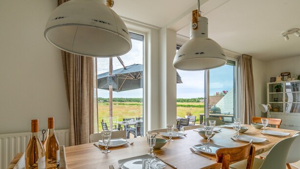 Ein Essbereich mit einem Holztisch, gedeckt für vier Personen, großen Pendelleuchten und Blick auf ein grünes Feld durch große Fenster.