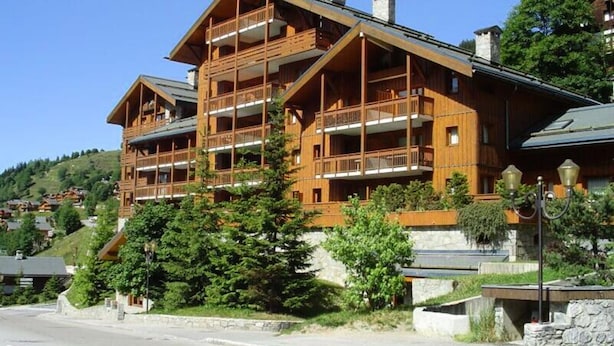 A multi-story wooden lodge with balconies, surrounded by greenery and set against a hilly backdrop. - Beautiful 3-room Duplex Apartment in the Center of Meribel, Close to Shops and ski Slopes (Les Allues)