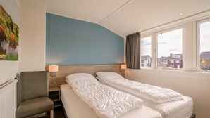 Et hotelværelse med to senge, en stol, et natbord, en lampe og et vindue med udsigt til bygninger.