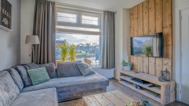 En moderne stue med en grå sofa, et træ-tv-skab og et stort vindue med gardiner.
