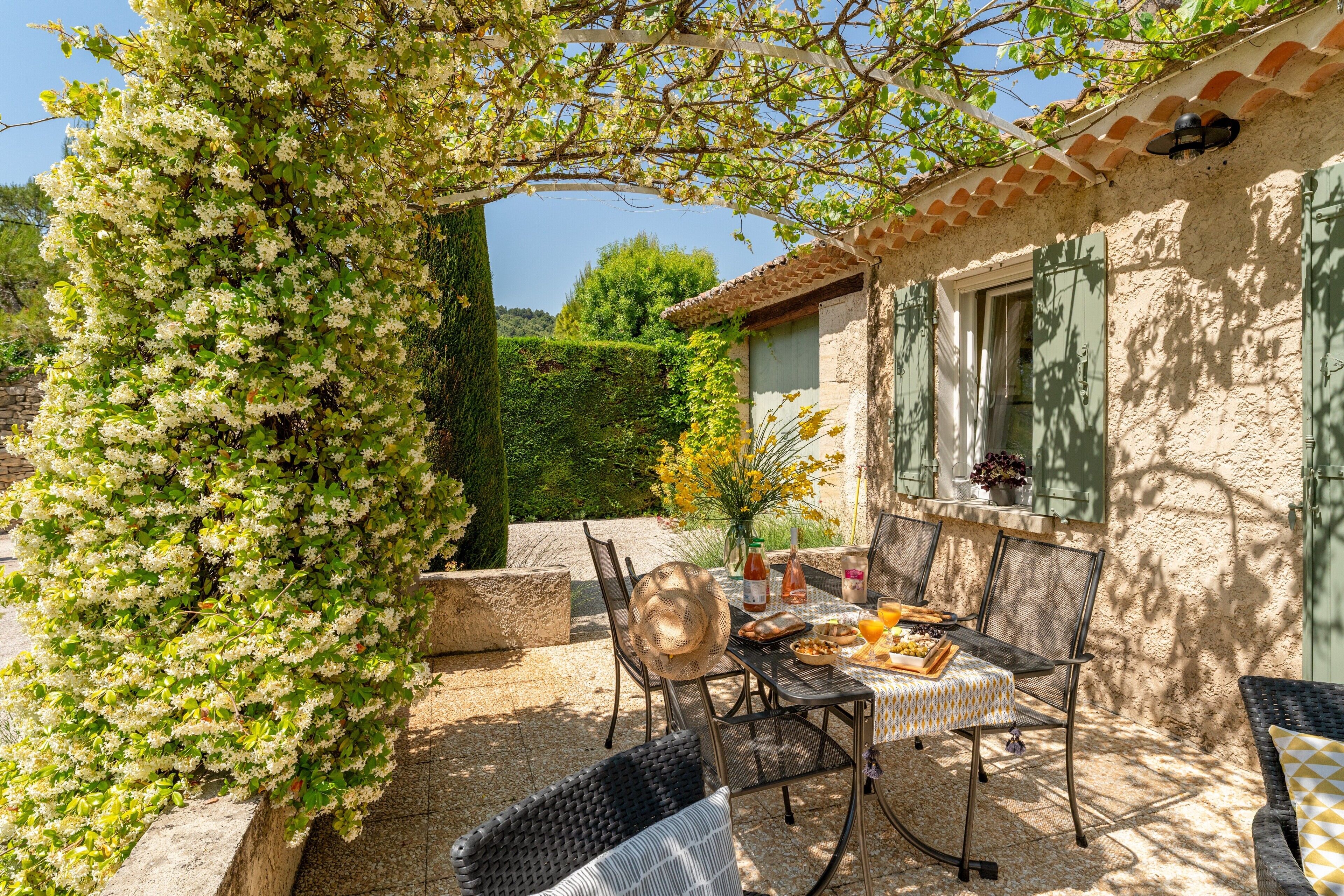 A patio with a table set for dining, surrounded by wicker chairs, under a pergola with climbing plants.
