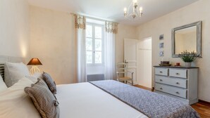 Ein Schlafzimmer mit Bett, Kommode, Spiegel, Stuhl und einem Fenster mit Vorhängen.