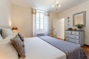 Ein Schlafzimmer mit Bett, Kommode, Spiegel, Stuhl und einem Fenster mit Vorhängen.