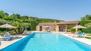 Ein Schwimmbecken mit Liegestühlen und Sonnenschirmen, ein Gebäude mit Ziegeldach und üppiges Grün im Hintergrund.