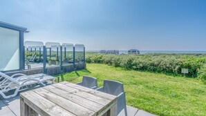 Un patio con mesa y sillas de madera, con vista a un área verde y una vista despejada del mar y edificios lejanos.