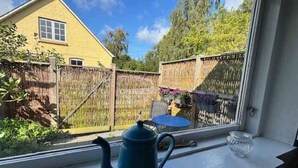 Blick aus einem Fenster auf eine Fensterbank mit einem blauen Teekessel, einen Holzzaun und ein Haus im Hintergrund.