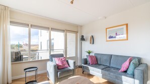 A modern living room with a grey sofa, armchairs, a coffee table, and a large window offering a view of the city. - 2 Person Island View Apartment (De Koog)
