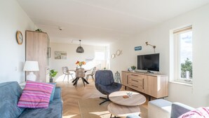 A modern living room with a sofa, armchair, coffee table, and a TV. There is a dining area in the background with a table and chairs. - 2 Person Island View Apartment (De Koog)