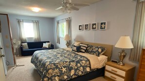 Quarto com cama, mesa de cabeceira, abajur, ventilador, sofá e parede com quadros.