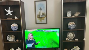 Uma televisão exibindo previsão do tempo, rodeada por prateleiras decorativas com diversos objetos.
