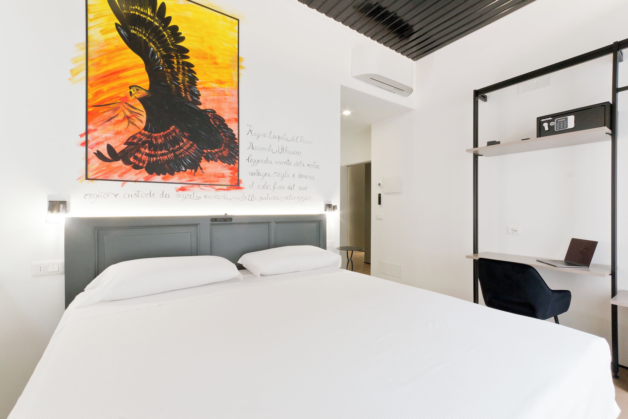 A hotel room with a large bed, a desk with a laptop, a chair, and a wall with a painting and text.