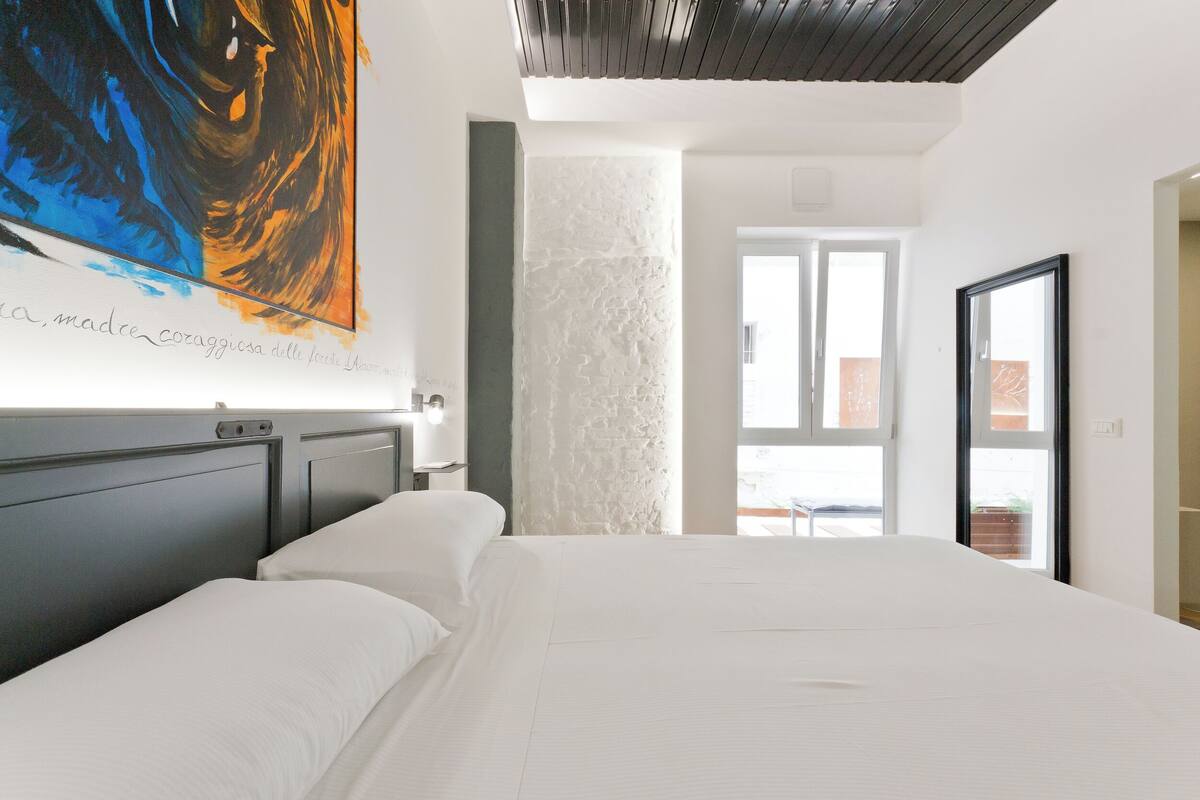 Ein modernes Schlafzimmer mit einem großen Bett, einem weißen Kopfteil, einem großen abstrakten Gemälde und Blick durch eine Glastür nach draußen.