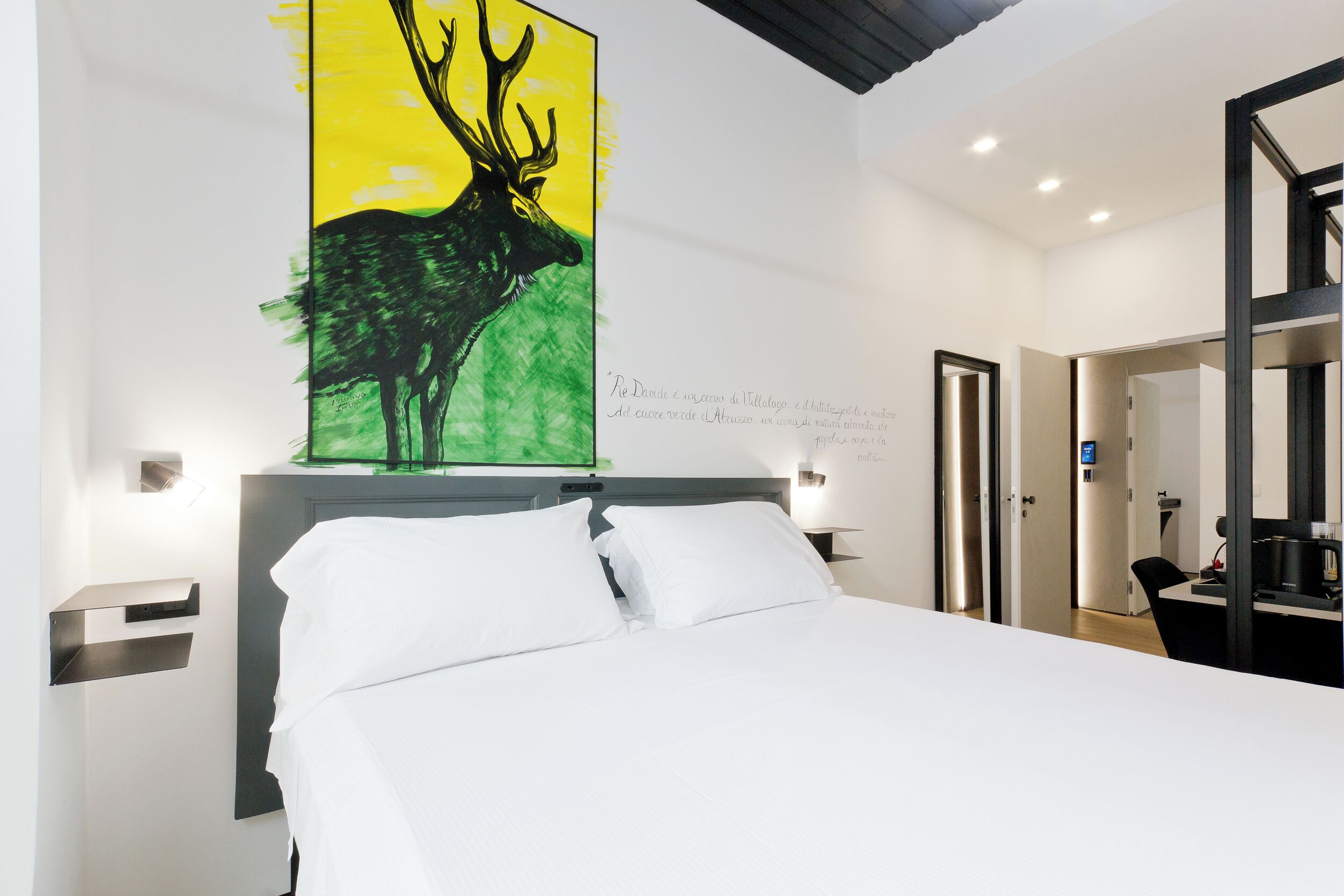 Una habitación de hotel con una cama, un cuadro de un ciervo, una mesita de noche, una lámpara de pared y una puerta que da a otra habitación.