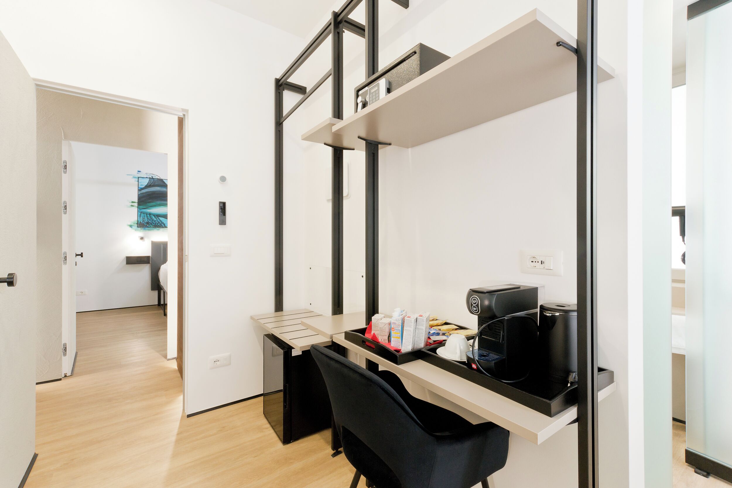Ein moderner Büroraum mit Schreibtisch, Stuhl und Kaffeemaschine. Über dem Schreibtisch befindet sich ein Regal und eine Tür zu einem weiteren Raum.