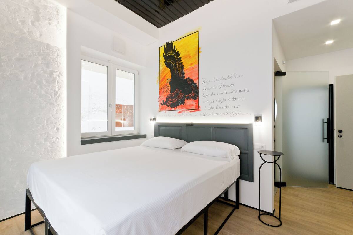 Ein modernes Schlafzimmer mit einem großen Bett, einem Fenster, einem Wandbild und einem kleinen Tisch.