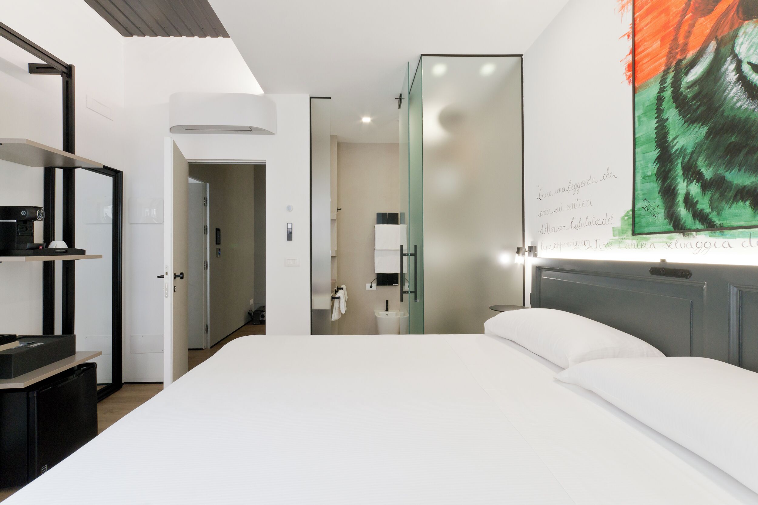 A modern hotel room with a large bed, a glass partition wall, and a wall-mounted artwork.