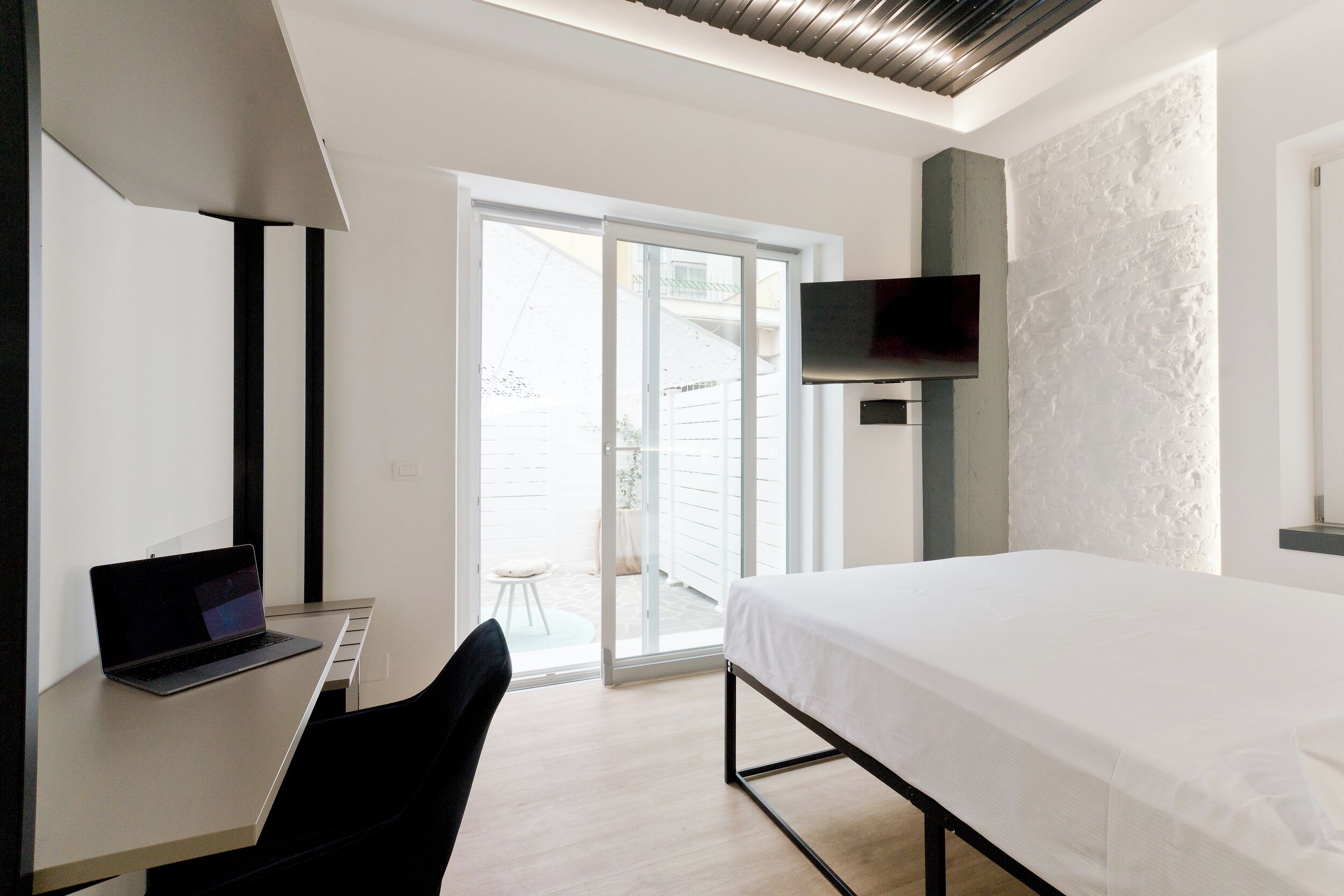 Une chambre d’hôtel moderne équipée d’un lit, d’un bureau avec un ordinateur portable, d’un téléviseur fixé au mur et d’une porte-fenêtre coulissante donnant sur un espace extérieur.