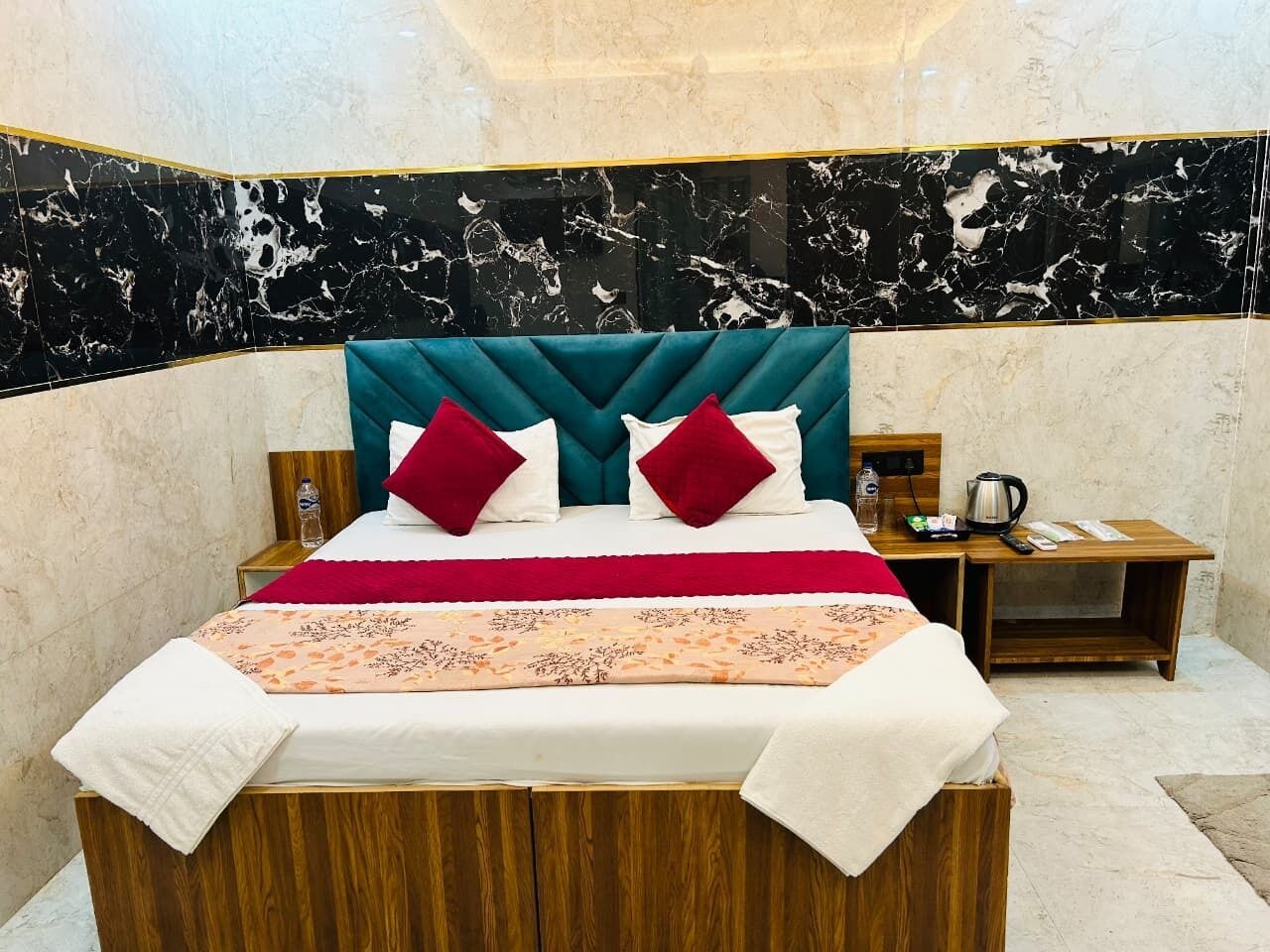 Una cama con cabecera color aguamarina, almohadas rojas y blancas y una colcha estampada con motivos florales. Junto a la cama, una mesita de noche de madera con una tetera y una botella de agua.