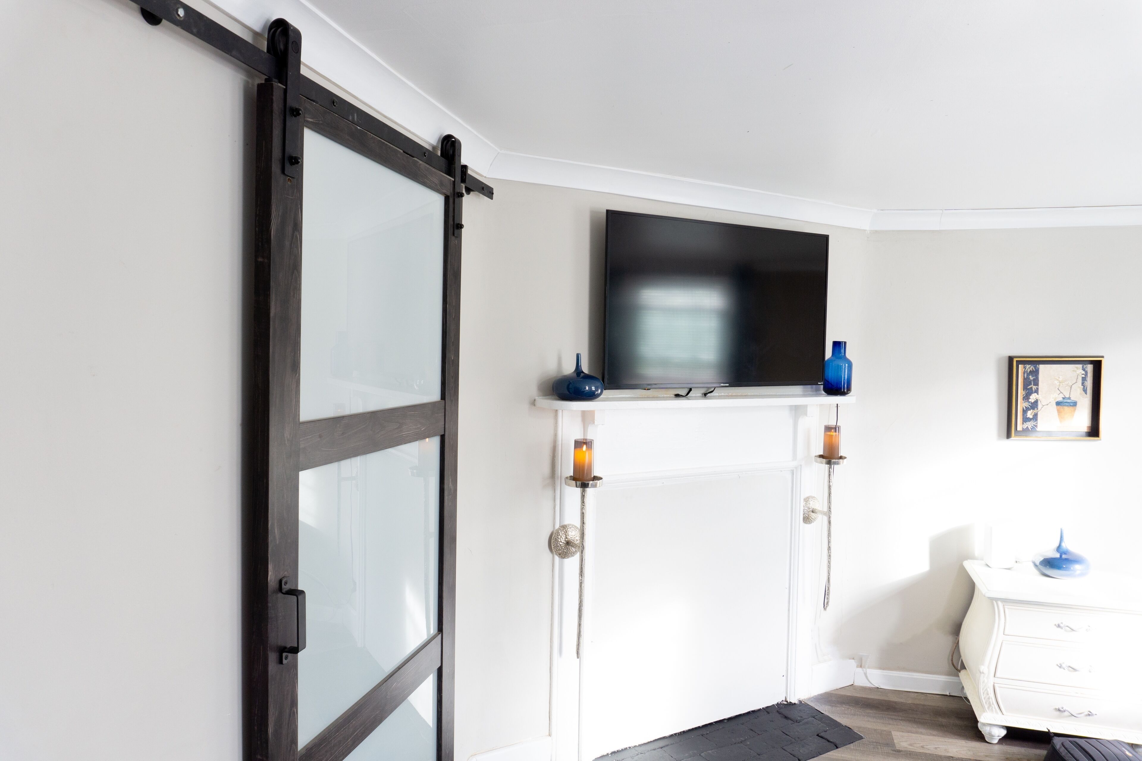Um quarto com porta deslizante, lareira e TV acima, e cômoda branca com um vaso azul.