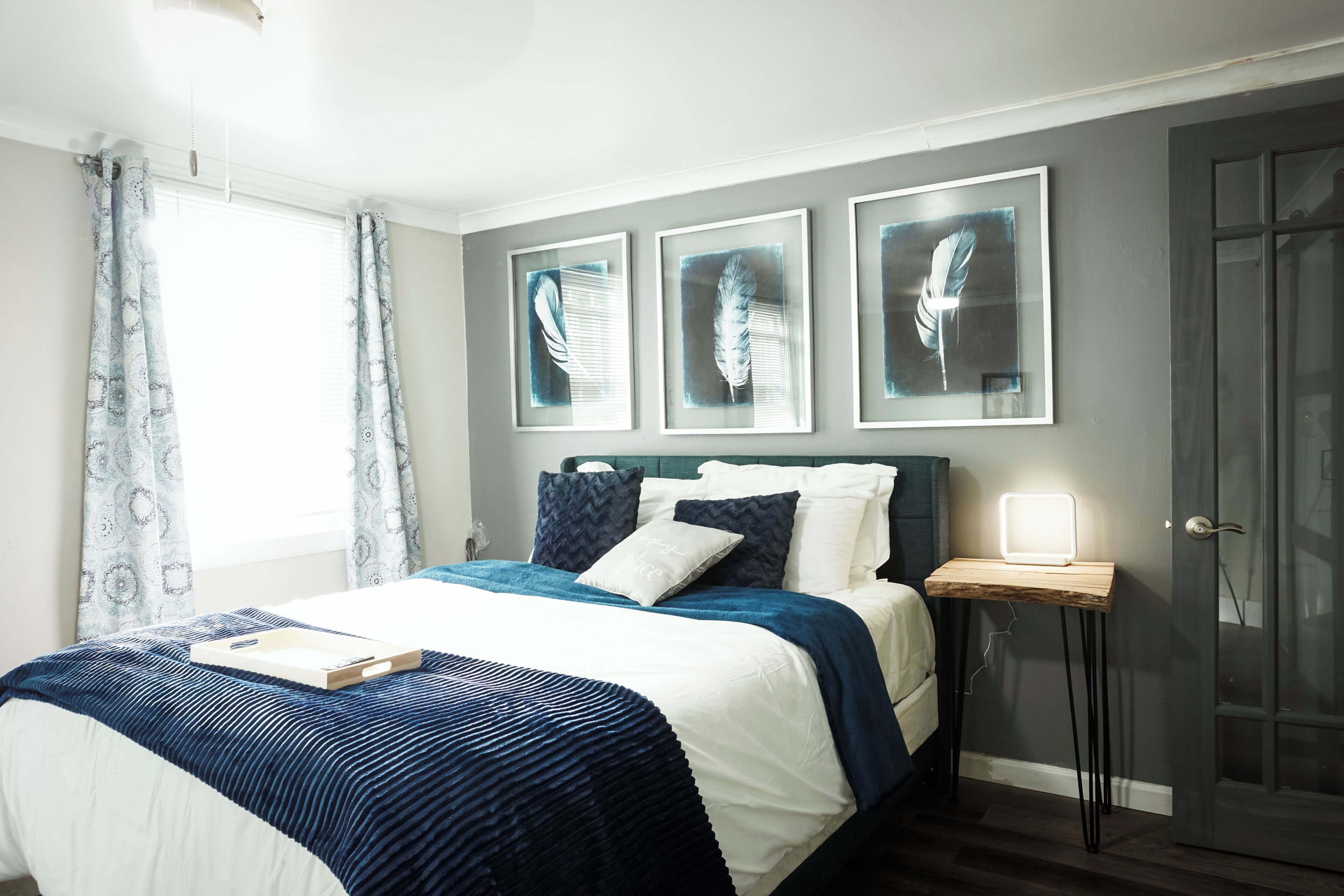 A neatly made bed with a blue and white bedspread, a bedside table with a lamp, and three framed artworks on the wall.