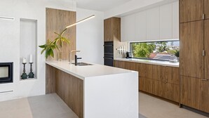 A modern kitchen with a central island, wooden cabinets, a built-in oven, and a window with a view of the outdoors. - Villa Verda 2 - Luxury Coastal Escape with Private Pool - Holiday Yamba (Yamba)