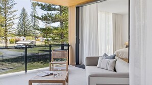 A balcony with a view of a parking area and trees, a sofa with cushions, and a table with glasses and a book. - Villa Verda 2 - Luxury Coastal Escape with Private Pool - Holiday Yamba (Yamba)