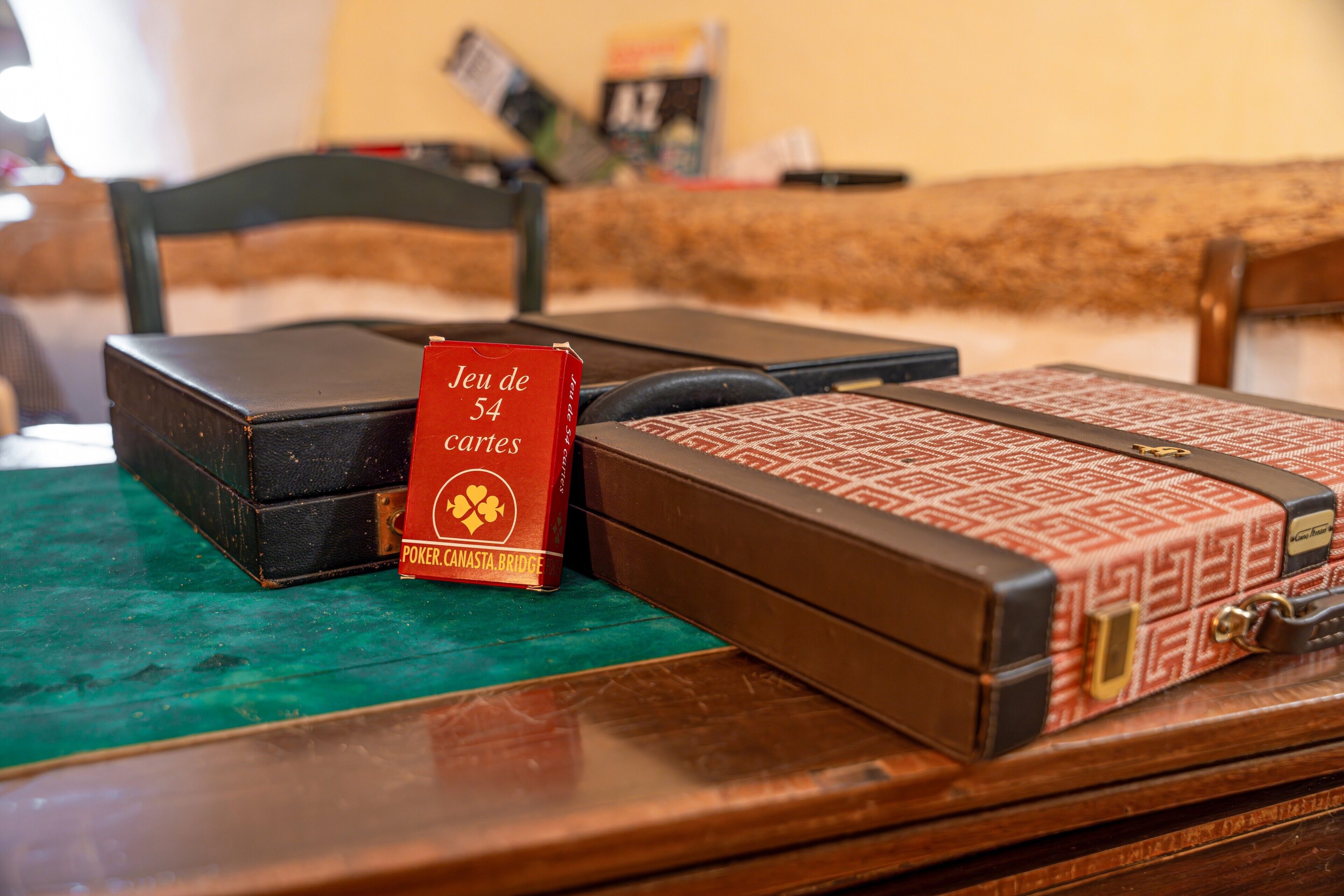 Ein Holztisch mit einer grünen Tischdecke, eine rote Schachtel mit dem Aufdruck 'Jeu de 54 cartes' und ein Musterkoffer.