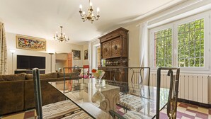 Een moderne woonkamer met een eettafel met glazen blad, een bank, een kroonluchter en een groot raam met uitzicht.