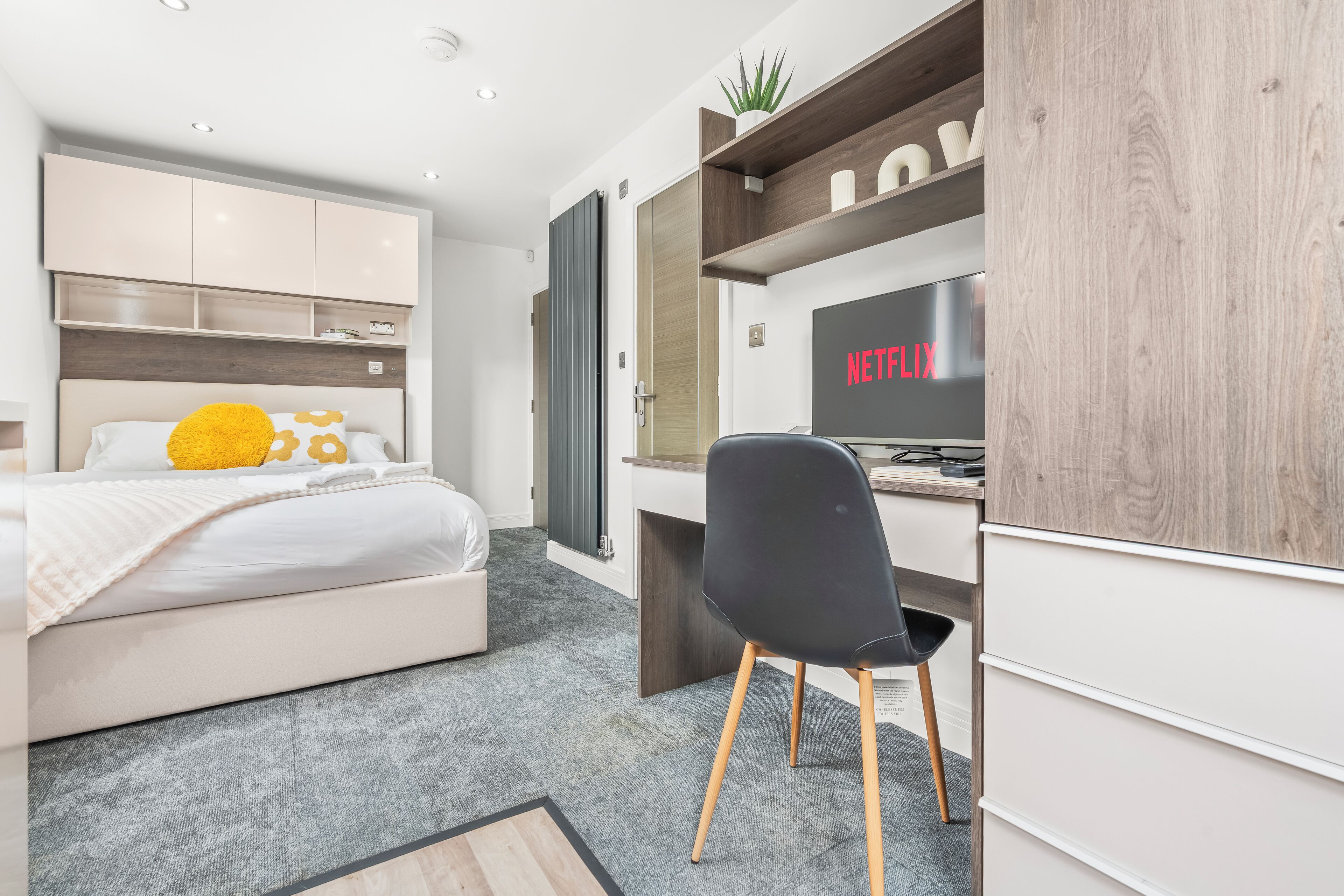 Ein modernes Schlafzimmer mit einem Bett, einem Schreibtisch, einem Sessel und einem Fernseher, der 'NETFLIX' anzeigt.