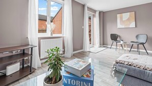 A modern living room with a large window, a radiator, a plant, a sofa, a coffee table, and a bookshelf. - Modern 4-Bed Close to Jaguar Whitley & City C… (Coventry)