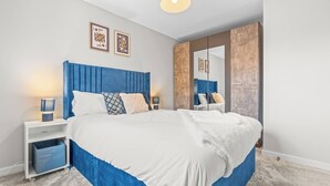 A modern bedroom with a large bed, a nightstand with a lamp, a wardrobe with a mirror, and two framed pictures on the wall. - Modern 4-Bed Close to Jaguar Whitley & City C… (Coventry)