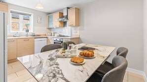 A modern kitchen with a marble countertop, wooden cabinets, and a dining area with a table set for two. - Modern 4-Bed Close to Jaguar Whitley & City C… (Coventry)