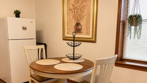 Un coin repas avec une table ronde en bois, deux chaises blanches, un réfrigérateur blanc, une estampe botanique encadrée au mur et une plante suspendue près de la fenêtre.