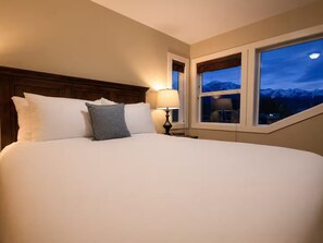 A neatly made bed with pillows, a bedside lamp, and a window with a mountain view. - Premier host-4BR/HotTub/Sauna/Massage Chairs (Revelstoke)