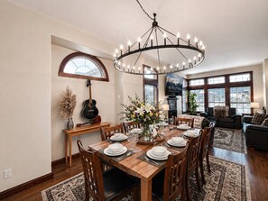 Un coin repas avec une table en bois dressée pour six personnes, entourée de chaises, d’un lustre et d’une guitare posée sur un support.