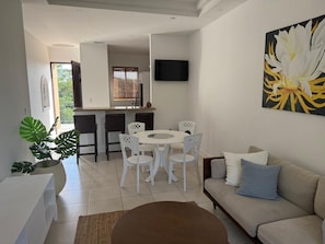 Uma sala de estar moderna com um sofá bege, uma mesa de centro e uma área de jantar com uma mesa redonda e cadeiras brancas.