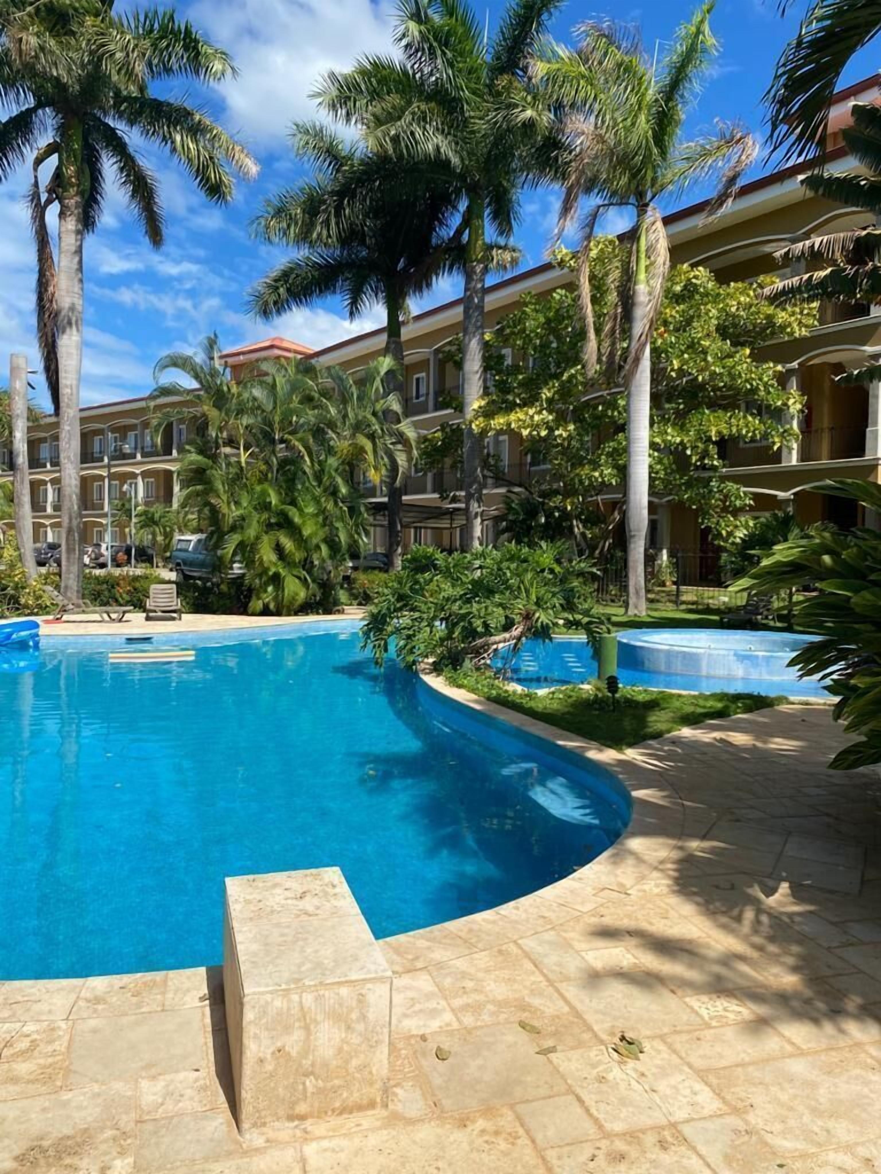 A swimming pool surrounded by palm trees and a multi-story building with balconies.