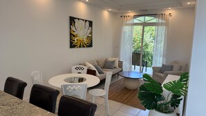 Una sala de estar moderna con comedor, que incluye un conjunto de sofá, una mesa de centro redonda y una mesa de comedor con sillas. Hay un ventanal con cortinas, un cuadro en la pared y una planta en maceta.