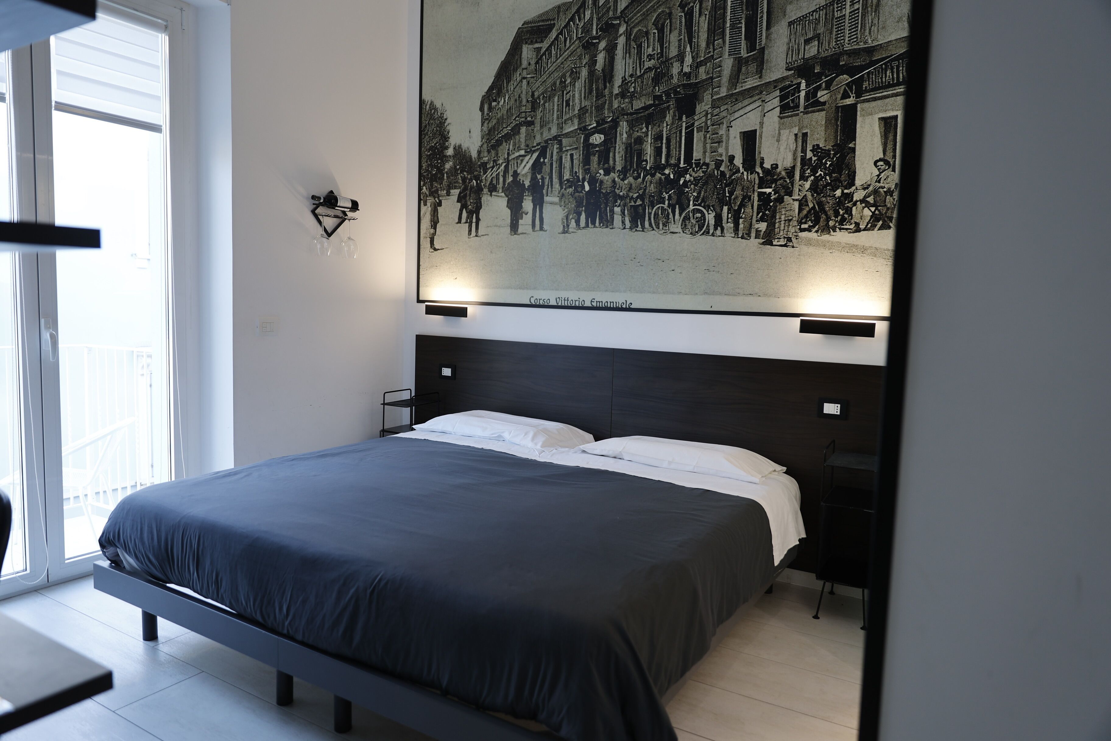 Un dormitorio moderno con una cama grande, una ventana con persianas y un cuadro colgado en la pared que muestra una escena de calle histórica.