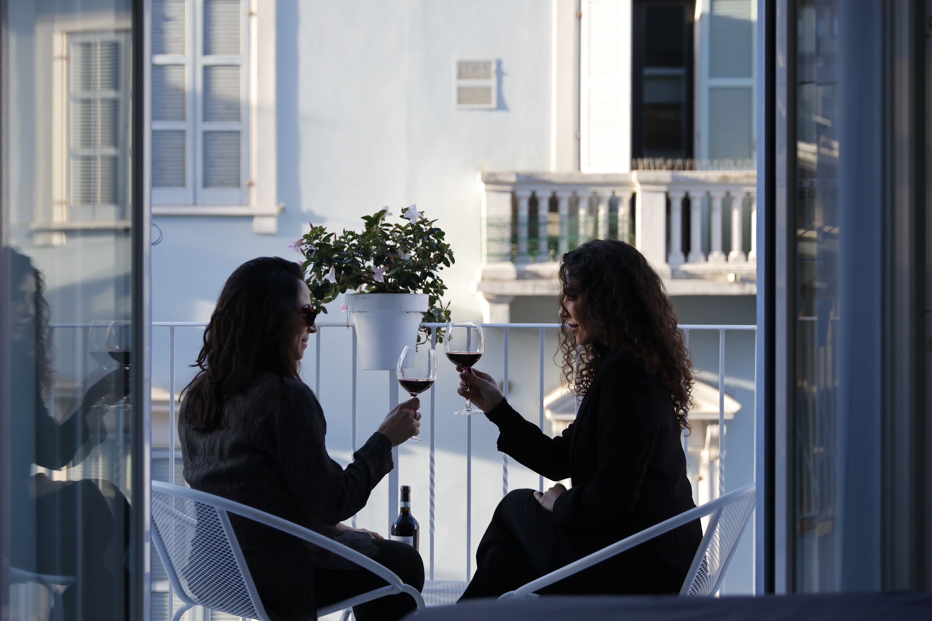 Dos personas sentadas en un balcón con una planta en maceta, sosteniendo copas de vino.