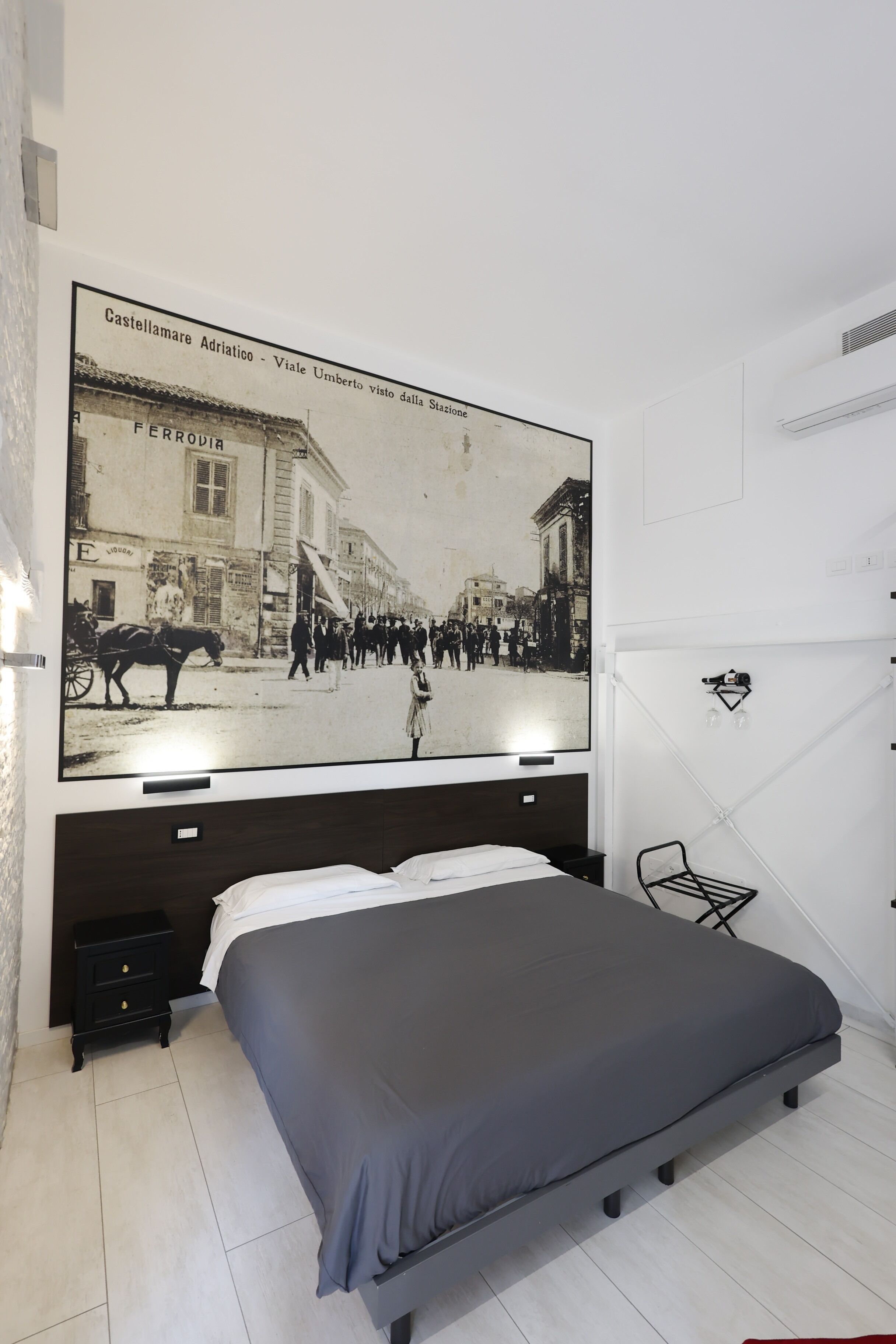 Een slaapkamer met een groot bed, een nachtkastje, een aan de muur bevestigde televisie waarop een historisch straatsbeeld te zien is, en een eenvoudige stoel.